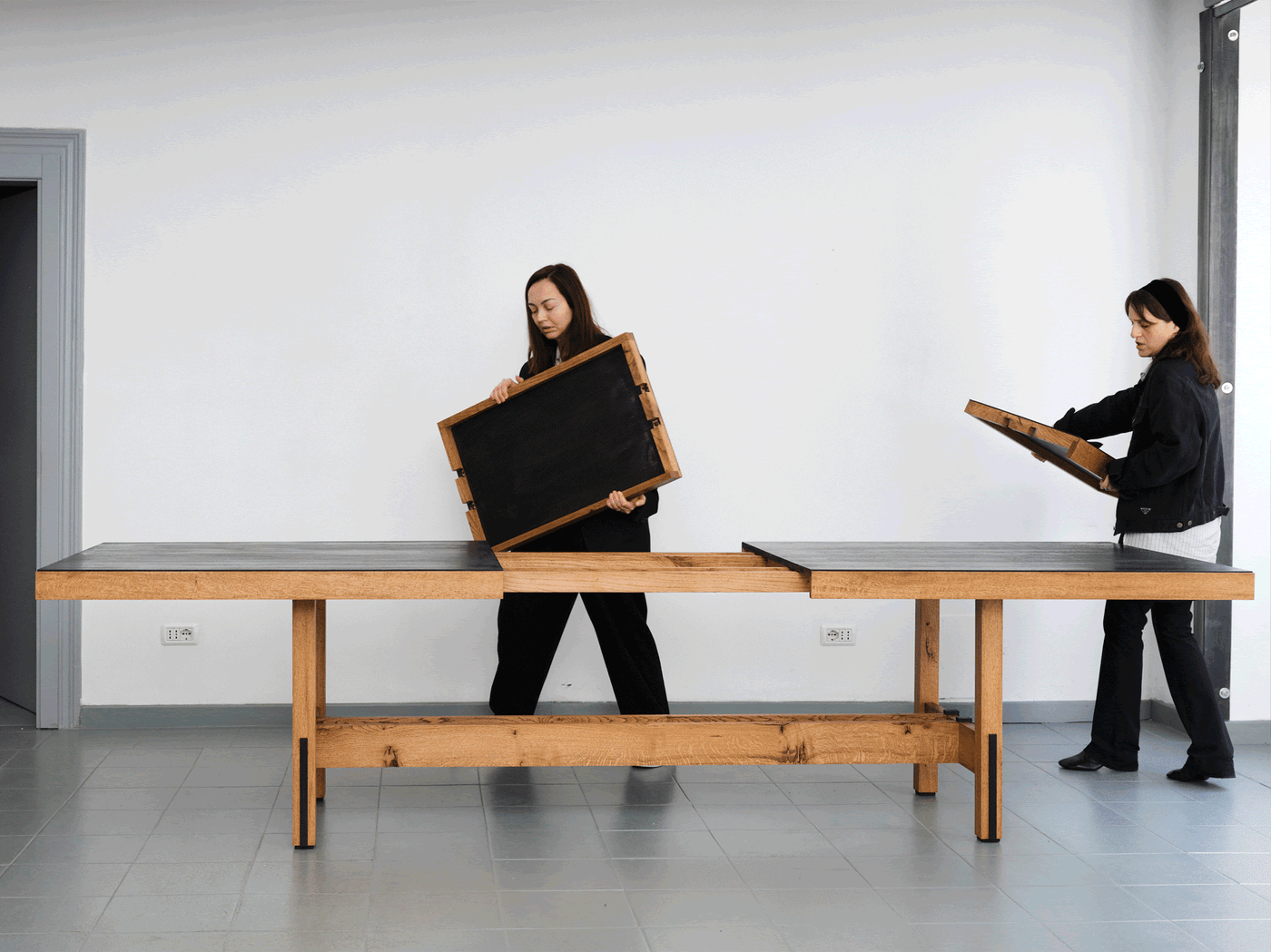 Nata Janberidze and Keti Toloraia of Rooms Studio interacting with a modular wooden table featuring removable black panels; the clean-lined piece combines natural oak with dark slate surfaces in a flexible, minimalist design setting against a white wall and tiled floor. Nata Janberidze and Keti Toloraia of Rooms Studio interacting with a modular wooden table featuring removable black panels; the clean-lined piece combines natural oak with dark slate surfaces in a flexible, minimalist design setting against a white wall and tiled floor.